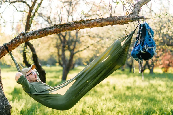 Heureux voyageur barbu hipster homme au chapeau vert couché en hummock sur pommier dans le jardin fleuri en été journée ensoleillée en vacances. Week-end et vacances. Tourisme. Loisirs. Rêver. Parfum dans l'air . — Photo