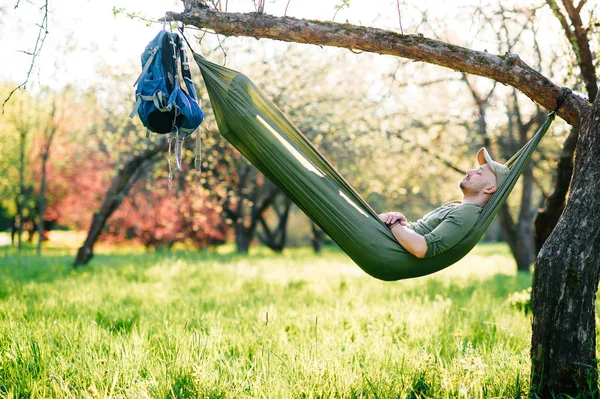 Feliz viajero barbudo hipster hombre en sombrero verde acostado en la burla en el manzano en el jardín floreciente en verano día soleado de vacaciones. Fin de semana y vacaciones. Turismo. Ocio. Soñando. Fragancia en el aire . — Foto de Stock