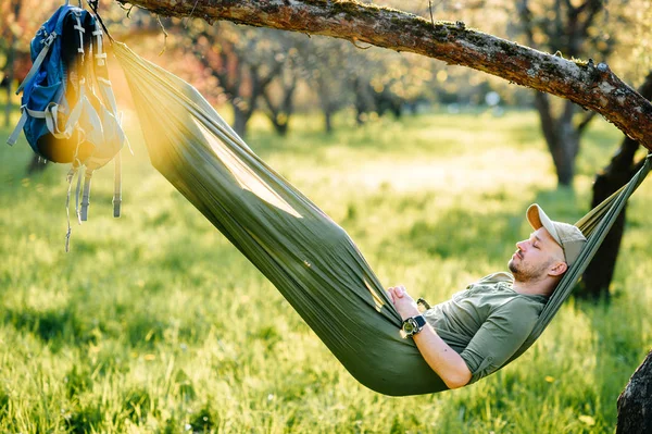 Heureux voyageur barbu hipster homme au chapeau vert couché en hummock sur pommier dans le jardin fleuri en été journée ensoleillée en vacances. Week-end et vacances. Tourisme. Loisirs. Rêver. Parfum dans l'air . — Photo