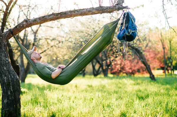 Heureux voyageur barbu hipster homme au chapeau vert couché en hummock sur pommier dans le jardin fleuri en été journée ensoleillée en vacances. Week-end et vacances. Tourisme. Loisirs. Rêver. Parfum dans l'air . — Photo