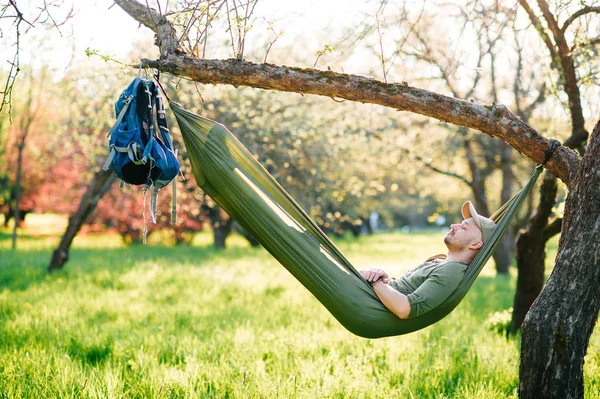 Счастливый путешественник бородатый хипстер человек в зеленой шляпе лежал в комок на яблоне в цветущем саду в летний солнечный день на отдыхе. Выходные и праздники. Туризм. Отдых. Мечтает. Аромат в воздухе . — стоковое фото
