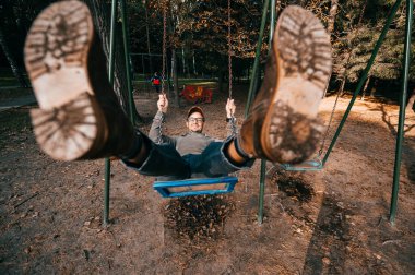Garip, tuhaf, tuhaf insanlar konsepti. Çocuk parkında salıncakta sallanan pahalı, modaya uygun botları olan yetişkin bir adam. Çok komik bir fikir. Çocuk içeride. Bacaklarını aç.. 
