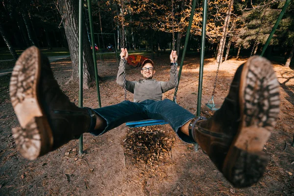 Strano bizzarro concetto di gente eccentrica. Uomo adulto in stivali alla moda cari d'annata che cavalcano su un'oscillazione in parco di città su parco giochi per bambini. Idea assurda e divertente. Bambino dentro. Gambe divaricate. — Foto Stock