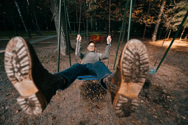 Extraño concepto excéntrico extraño de la gente. Hombre adulto en botas de moda costosas vintage cabalgando en un columpio en el parque de la ciudad en el patio de recreo para niños. Qué idea más graciosa. Niño dentro. Las piernas extendidas. — Foto de Stock