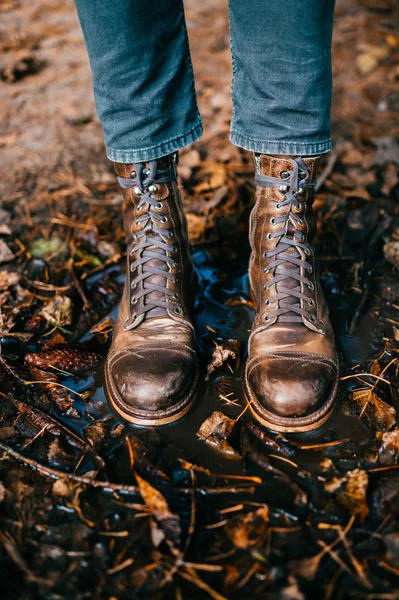 Close-up van de benen van de mens in fashinable trendy Italiaanse hipster vintage oude gekrast laarzen staan in plas. Natte en rauwe herfst. De herfstkleuren. Sinaasappelbladeren, dennenappels in het water. Vreemd bizar concept.. — Stockfoto