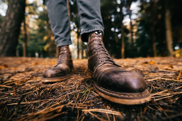 Мужские ноги в винтажных кожаных сапогах и обрезанных джинсах стоят в осеннем лесу на земле. Осенние цвета и концепция настроения. Удобная обувь. Прогулки на выходные на природе. Дисторсия объектива. — стоковое фото