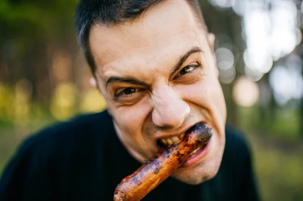 Closeup portrait of male licking and biting roasted sausage with disgusting ugly monstrous facial expression. Meat lover. Sinister evil diabolical face. Bum with maniacal eyes. Depth of filed. Funny.