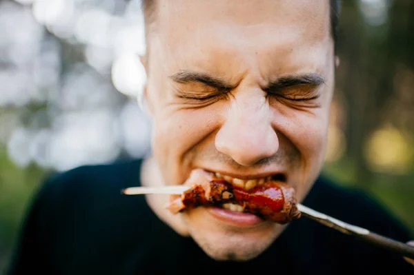 Facial expression of man eating with disgust grilled sausage outdoor. Vegetarian feeling pain and suffering trying meat. Pork is bad. Recycled dead animal. Tasteless fried food. Adult male biting. — Stock Photo, Image