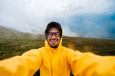 Selfie portrait of a smiling and laughing traveller man in yellow raincoat and glasses in the clouds mountains in stromy weather with rain. clipart