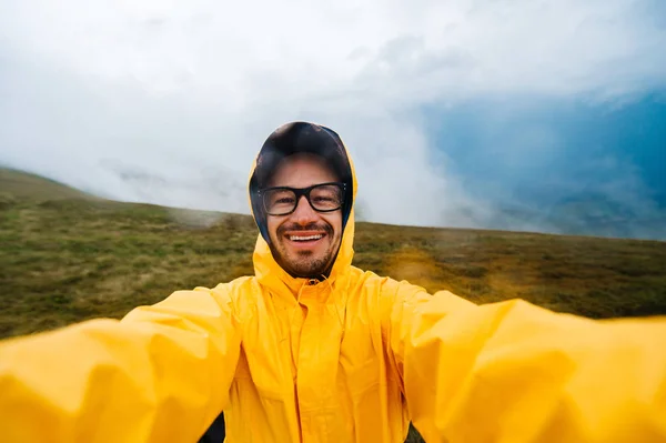 Selfie retrato de um homem viajante sorridente e rindo em capa de chuva amarela e óculos nas nuvens montanhas em tempo estropiante com chuva. — Fotografia de Stock