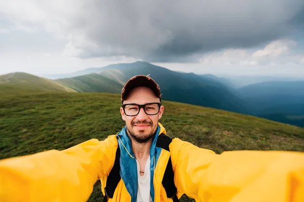 Retrato selfie de un viajero sonriente y risueño con impermeable amarillo y gafas en las nubes montañas en un clima caluroso con lluvia. — Foto de Stock