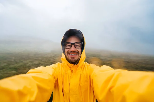 Retrato selfie de un viajero sonriente y risueño con impermeable amarillo y gafas en las nubes montañas en un clima caluroso con lluvia. —  Fotos de Stock