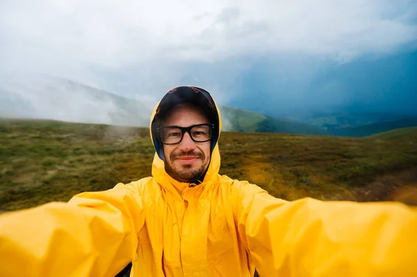 Selfie retrato de um homem viajante sorridente e rindo em capa de chuva amarela e óculos nas nuvens montanhas em tempo estropiante com chuva. — Fotografia de Stock