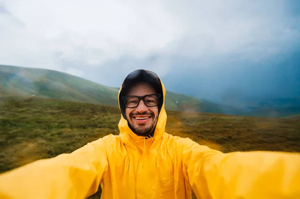 Retrato selfie de un viajero sonriente y risueño con impermeable amarillo y gafas en las nubes montañas en un clima caluroso con lluvia. — Foto de Stock