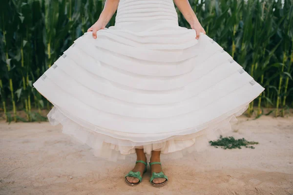 Одинокая неузнаваемая молодая счастливая невеста, стоящая в белом красивом платье после свадебной церемонии. Празднование свадьбы. Женщина на дороге возле огуречного поля показывает грязные пыльные сандалии. Веселая концепция . — стоковое фото