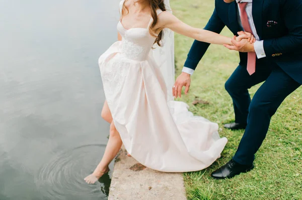 Novia irreconocible en vestido de novia elegante caro empujando su pierna hermosa desnuda delgada en el río. novio desconocido en traje sosteniendo su esposa de la mano y el cuerpo. Celebración del día de boda. Caminata nupcial . —  Fotos de Stock