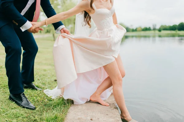 Unrecognizable bride in expensive stylish wedding dress pushing her slim naked beautiful leg in river. Unknown groom in suit holding his wife hand and body.  Wedding day celebration. Bridal walking. — Stock Photo, Image