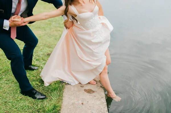 Novia irreconocible en vestido de novia elegante caro empujando su pierna hermosa desnuda delgada en el río. novio desconocido en traje sosteniendo su esposa de la mano y el cuerpo. Celebración del día de boda. Caminata nupcial . —  Fotos de Stock