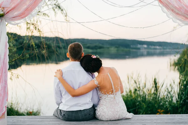 Feliz novia y novio después de la ceremonia sentado en el lugar de madera para el arco de la boda con decoración y flores. Pétalos de rosa y césped. Un par de amantes abrazándose al atardecer cerca del lago. Cabeza sobre hombro. Romántico . —  Fotos de Stock