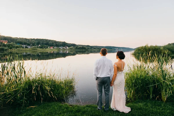 Feliz novia y novio después de la ceremonia de boda de pie en la orilla del lago por la noche al atardecer en la naturaleza. Una joven pareja casada tomando las manos y mirando al horizonte. Vista romántica. Celebración del matrimonio . —  Fotos de Stock