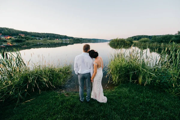 Feliz novia y novio después de la ceremonia de boda de pie en la orilla del lago por la noche al atardecer en la naturaleza. Una joven pareja casada tomando las manos y mirando al horizonte. Vista romántica. Celebración del matrimonio . —  Fotos de Stock