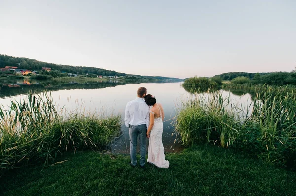 Feliz novia y novio después de la ceremonia de boda de pie en la orilla del lago por la noche al atardecer en la naturaleza. Una joven pareja casada tomando las manos y mirando al horizonte. Vista romántica. Celebración del matrimonio . —  Fotos de Stock