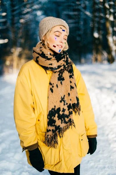 En ganska ung udda flicka med änglavackra dockögon. bisarr kvinna i varma kläder stående på vintern snöig skog utomhus. Fin blond tonåring härlig ansikte med färgglada klistermärken i ansiktet. — Stockfoto