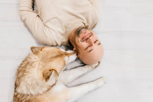 Взрослый лысый мужчина с хаски щенком спит на полу. Оунер с домашним питомцем. Добрые и душевные эмоции. Прекрасная собака отдыхает с молодым мужчиной. Парень с любимым домашним животным, обнимающим друг друга. — стоковое фото
