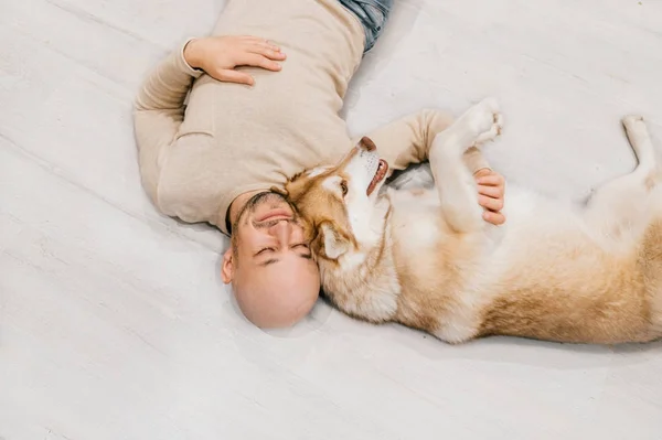 Ein erwachsener Mann mit Glatze und Husky-Welpen schläft auf dem Boden. Besitzer mit Haustier zusammen zu Hause. Freundliche und gefühlvolle Gefühle. Schöner Hund, der mit jungen Rüden ruht. Kerl mit geliebtem Haustier umarmt sich. — Stockfoto