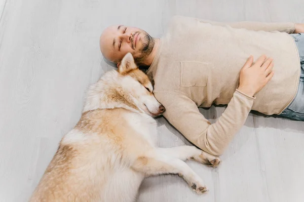 Uomo adulto calvo con cucciolo husky che dorme sul pavimento. Proprietario con animale domestico insieme a casa. Emozioni gentili e sentimentali. Bel cane a riposo con giovane maschio. Ragazzo con amato animale domestico che si abbraccia. — Foto Stock