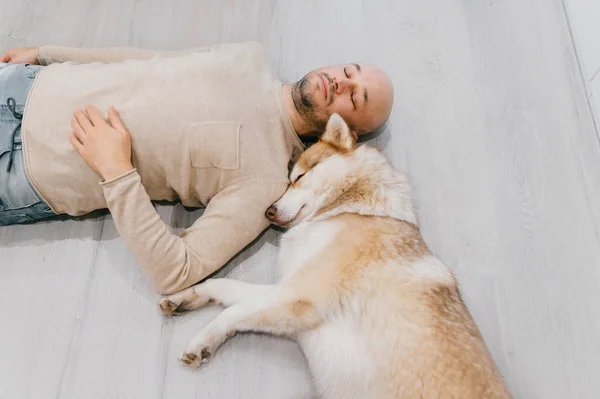 Dospělý plešatý muž s husky štěně spí na podlaze. Majitel s domácím mazlíčkem. Milé a oduševnělé emoce. Krásný pes odpočívající s mladým samcem. Chlap s milovaným domácím zvířetem objímající se navzájem. — Stock fotografie