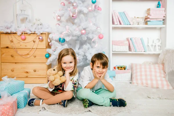 Kinderen zitten op de vloer op tapijt met Nieuwjaars versieringen en kerstboom. Emotionele kinderen gezichten. Knappe jongen en prinses meisje camera kijken. Gelukkige mensen samen op vakantie — Stockfoto