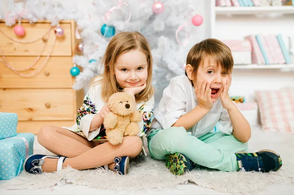Niños sentados en el suelo en la alfombra con decoraciones de año nuevo y árbol de Navidad. Caras de niños emocionales. Niño guapo y niña princesa mirando a la cámara. Gente feliz juntos en vacaciones —  Fotos de Stock