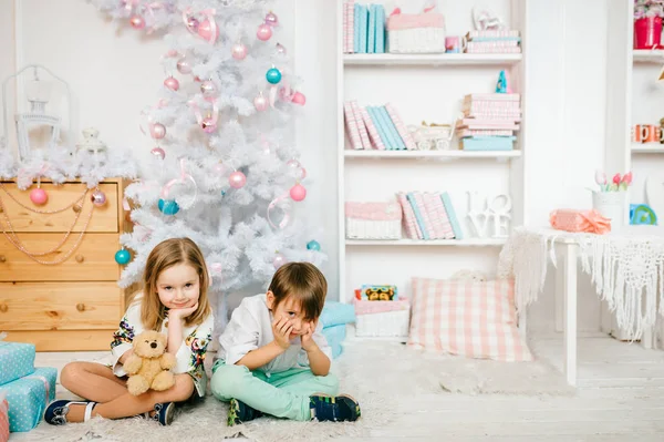 Niños sentados en el suelo en la alfombra con decoraciones de año nuevo y árbol de Navidad. Caras de niños emocionales. Niño guapo y niña princesa mirando a la cámara. Gente feliz juntos en vacaciones —  Fotos de Stock