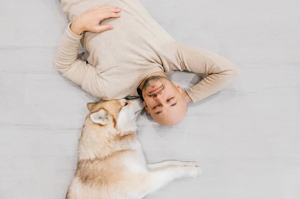 Ein erwachsener Mann mit Glatze und Husky-Welpen schläft auf dem Boden. Besitzer mit Haustier zusammen zu Hause. Freundliche und gefühlvolle Gefühle. Schöner Hund, der mit jungen Rüden ruht. Kerl mit geliebtem Haustier umarmt sich. — Stockfoto