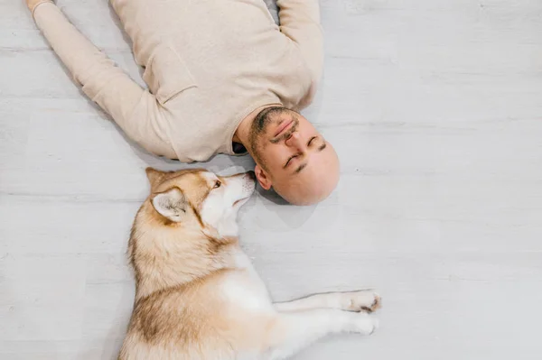Adult bald man with husky puppy sleeping on floor. Owner with pet together at home. Kind and soulful emotions. Lovely dog resting with young male. Guy with beloved domestic animal hugging each other.