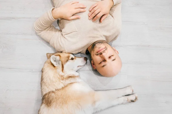 Ein erwachsener Mann mit Glatze und Husky-Welpen schläft auf dem Boden. Besitzer mit Haustier zusammen zu Hause. Freundliche und gefühlvolle Gefühle. Schöner Hund, der mit jungen Rüden ruht. Kerl mit geliebtem Haustier umarmt sich. — Stockfoto