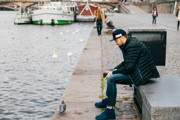 Lonely man sitting on bench in depression and thinking of problems in life. Abstract city background. Dove walking the embankment river in Prague. Psychological male portrait. Solitude and loneliness. — Stock Photo, Image