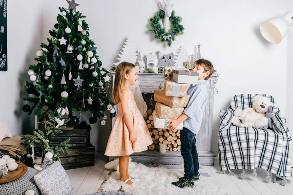 Fiatal jóképű fiú bemutatja új évet ajándékok a nevető aranyos kislány gyönyörű ruha. Karácsonyfa és ünnepi dekoráció. Szép pár vicces érzelmi gyermekek. Őszinte gyerekek otthon. — Stock Fotó