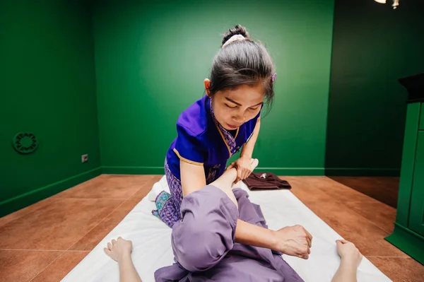 Young Thai masseur trainer girl in oriental exotic dress making and demonstrating different traditional spa ceremony procedures to european beautiful woman in pajamas in green relaxation yoga studio.