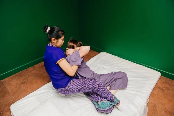 Young Thai masseur trainer girl in oriental exotic dress making and demonstrating different traditional spa ceremony procedures to european beautiful woman in pajamas in green relaxation yoga studio.