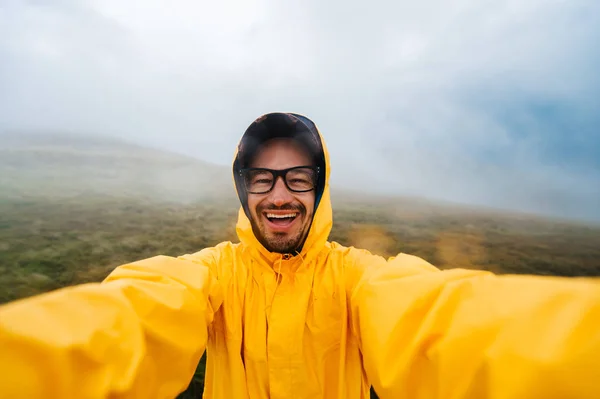 Retrato selfie de un viajero sonriente y risueño con impermeable amarillo y gafas en las nubes montañas en un clima caluroso con lluvia —  Fotos de Stock