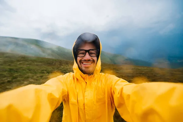 Retrato selfie de un viajero sonriente y risueño con impermeable amarillo y gafas en las nubes montañas en un clima caluroso con lluvia — Foto de Stock