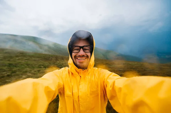 Selfie πορτρέτο ενός χαμογελαστού και γελαστού ταξιδιώτη με κίτρινο αδιάβροχο και γυαλιά στα σύννεφα βουνά σε λείο καιρό με βροχή — Φωτογραφία Αρχείου