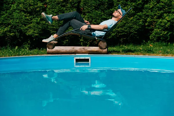 Uomo adulto che si rilassa in chaise longue oltre la piscina in villa in Sicilia con muro di alberi verdi meridionali sullo sfondo. Un ragazzo che prende il sole in vacanza. Tempo libero, spa e bella vita. — Foto Stock