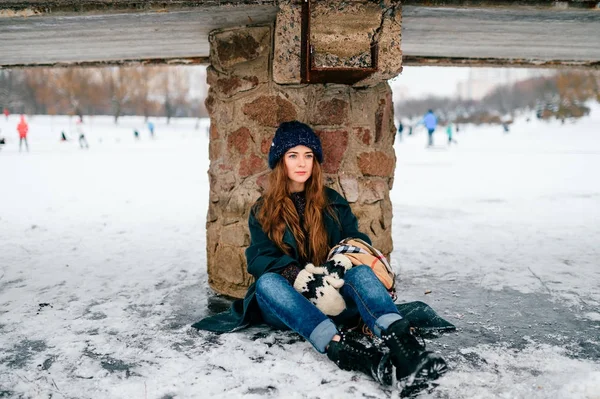 Joven chica adolescente hermosa de moda en ropa elegante con el pelo largo marrón sentado debajo del puente en el hielo en el lago congelado en heladas frías el día de diciembre de invierno en el parque de la ciudad. Mujer cansada y sin amigos . — Foto de Stock