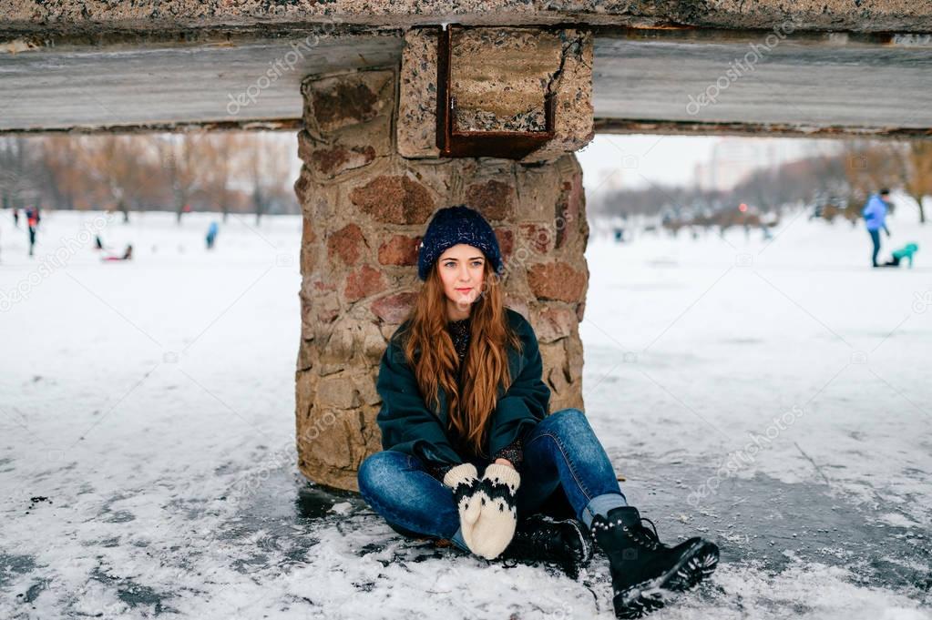 Young fashionable beautiful teenager girl in stylish clothes with long brown hair sitting under bridge on ice on frozen lake in cold frost winter december day in city park. Tired and friendless woman.