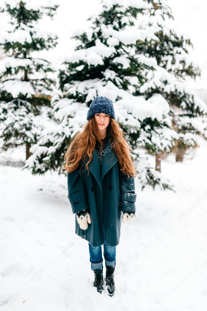 Young beautiful teenager girl in oversized wool coat with long brown beautiful hair standing in winter city park with snowy spruces on background. Fashionable stylish lady with amazaing blue eyes.