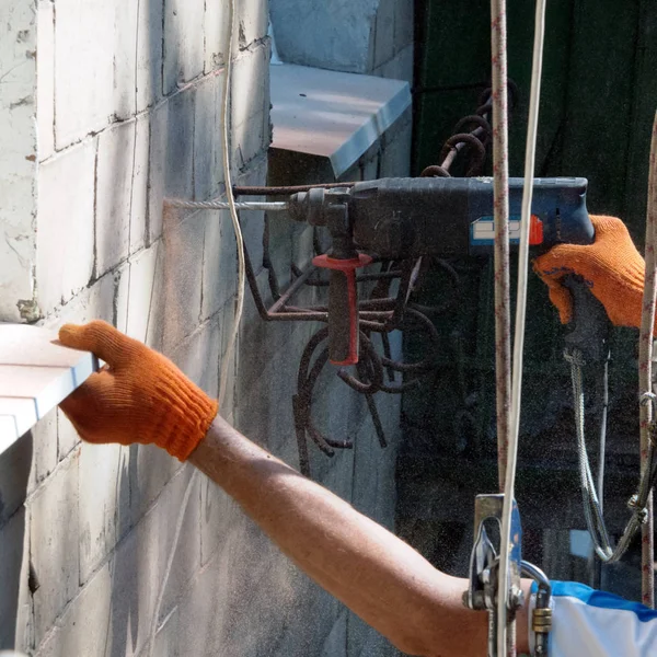 Aşağı boru kurulum süreci. Bir işçi, çok katlı binanın duvarını elektrikli matkapla deliyor. Her yere taş tozu saçılıyor..