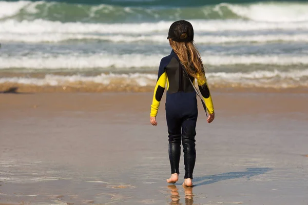 Menina em um boné e um terno de surfista está de pé na praia — Fotografia de Stock